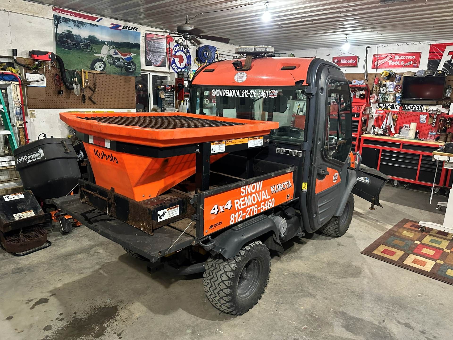 Orange UTV with a salt spreader attachment parked in a garage, labeled for snow removal services.