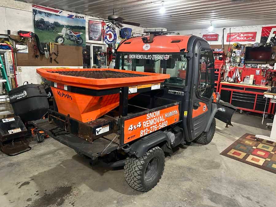 An orange salt spreader mounted on the back of a UTV parked inside a workshop with various tools and equipment.