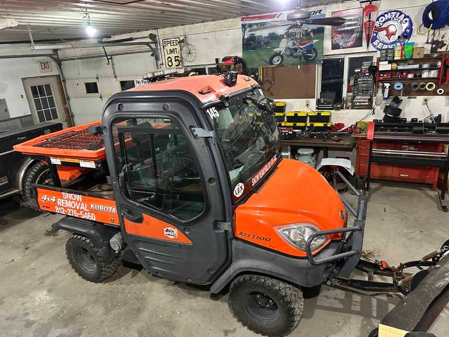 An orange Kubota RTV utility vehicle with a snowplow attachment, parked inside a garage.