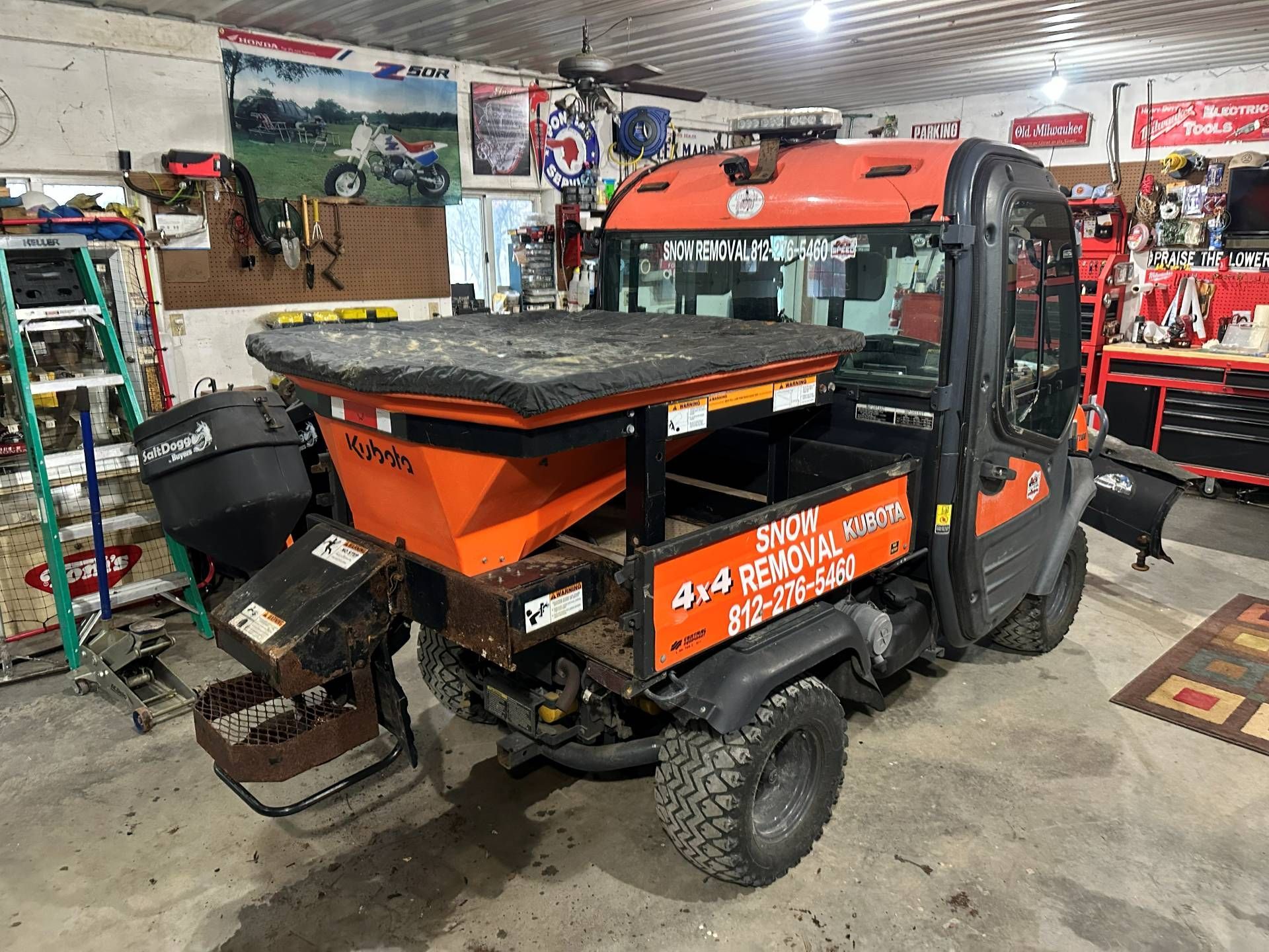 An orange, enclosed utility vehicle with a salt spreader attachment parked inside a cluttered workshop.