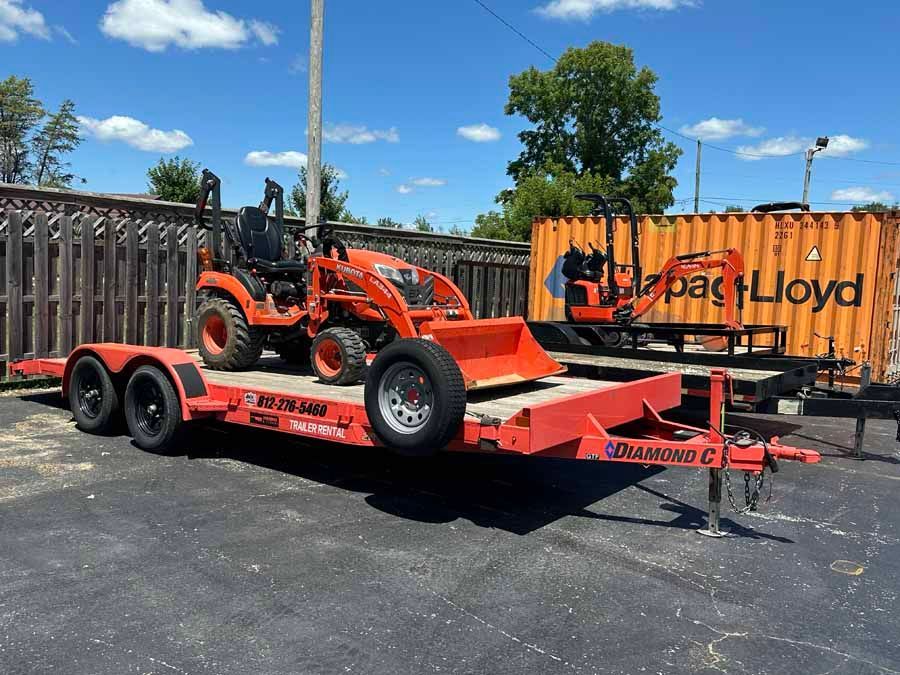 An orange tractor loader sits on a red flatbed trailer parked outside on a sunny day near an orange shipping container.