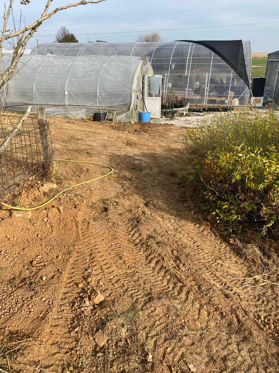 Two hoop-style greenhouses sit in a dirt lot with tire tracks, partially shaded by a black tarp on a sunny day.