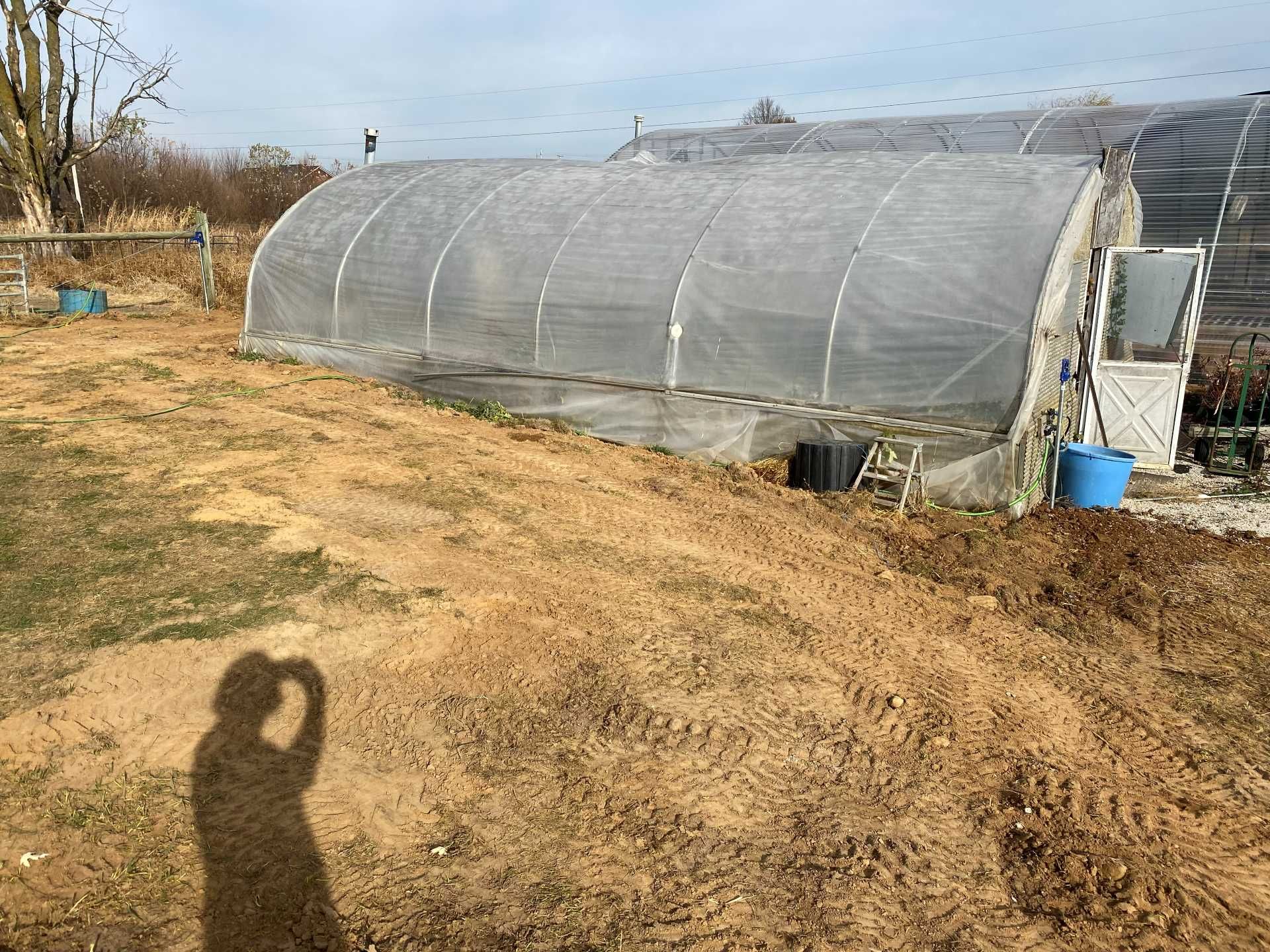 A long, tunnel-shaped greenhouse covered in translucent white plastic, situated on a plot of bare, light brown soil.