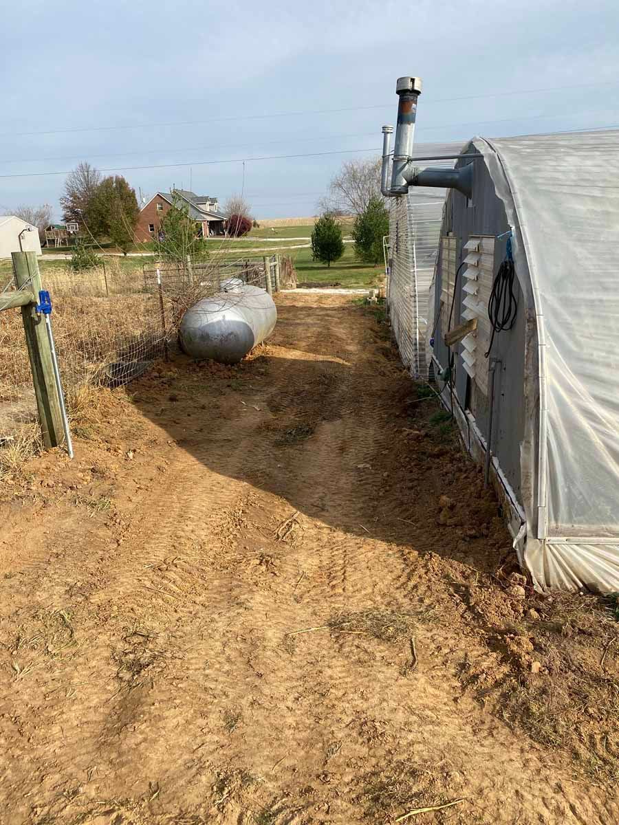 A dirt path runs between a greenhouse with a chimney and a propane tank in a rural setting.