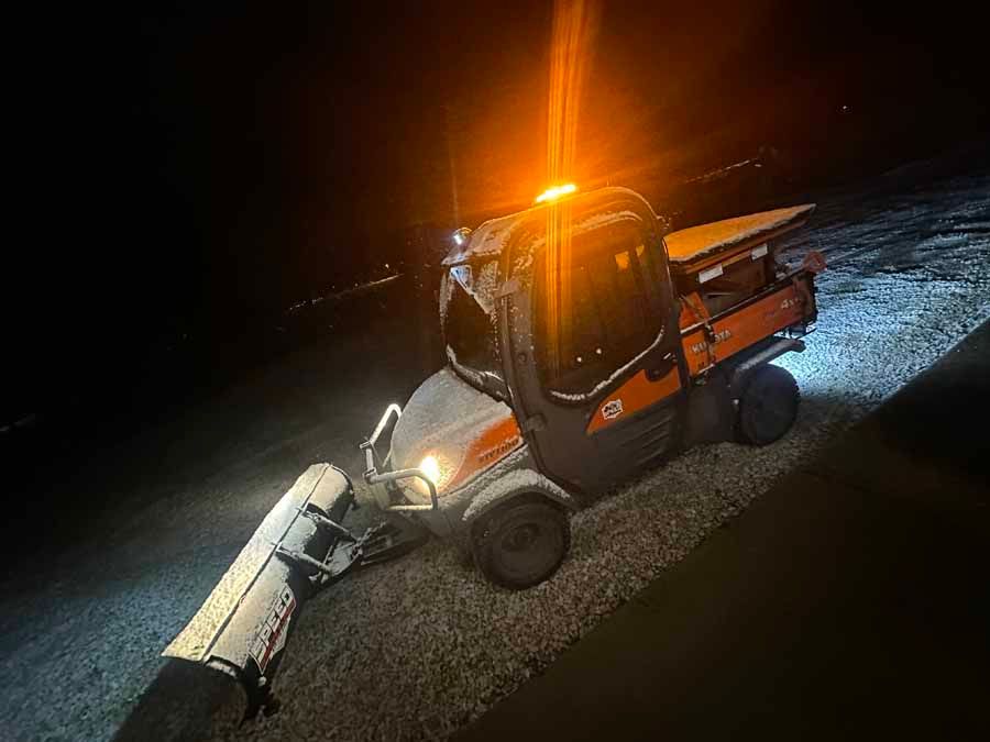 An orange utility vehicle with a snow plow attachment at night, featuring a bright, activated amber safety light.