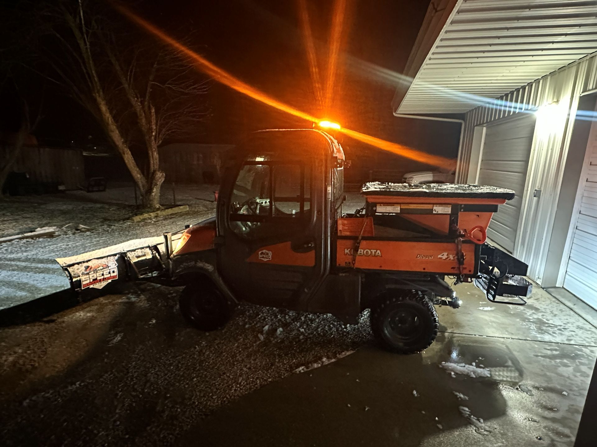 An orange Kubota utility vehicle with a snowplow and salt spreader parked on a snowy driveway at night.