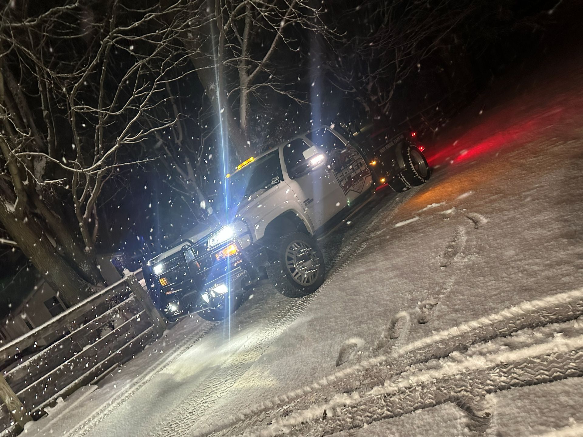 A white dually pickup truck parked at night on a snow-covered surface with its headlights illuminated during a snowfall.