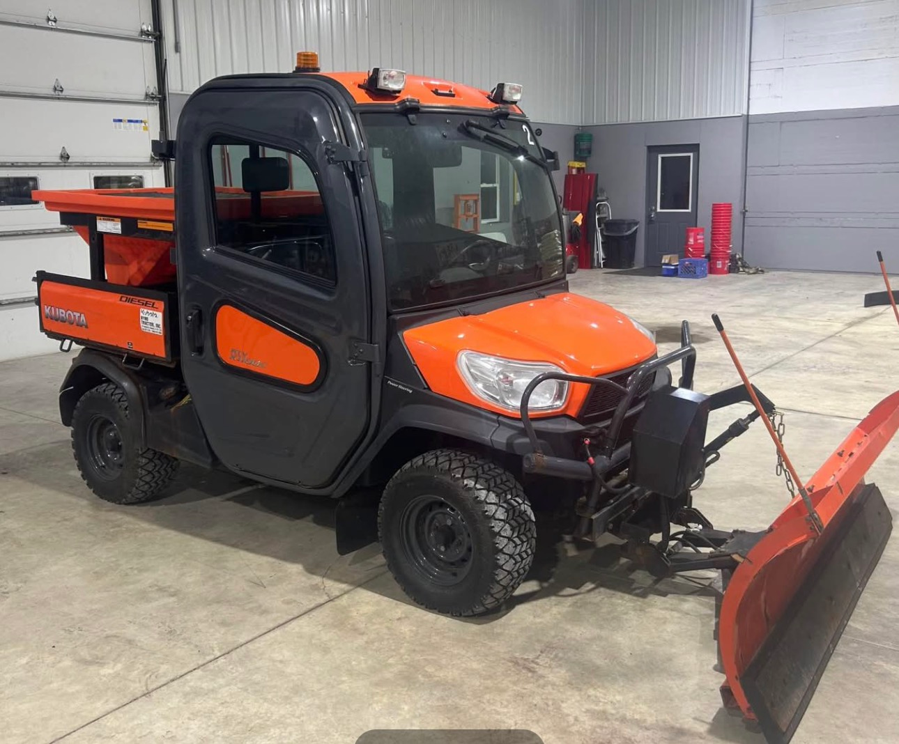 Orange Kubota utility vehicle equipped with a snow plow and a salt spreader inside a garage.