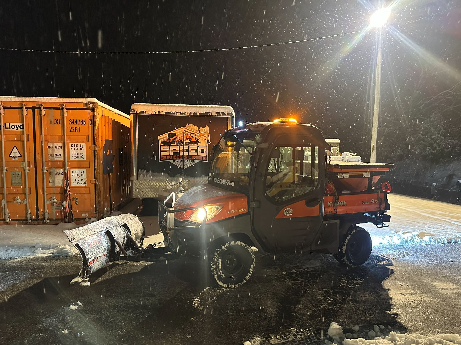 An orange utility vehicle with a snowplow parked at night in a snowy lot near orange shipping containers.
