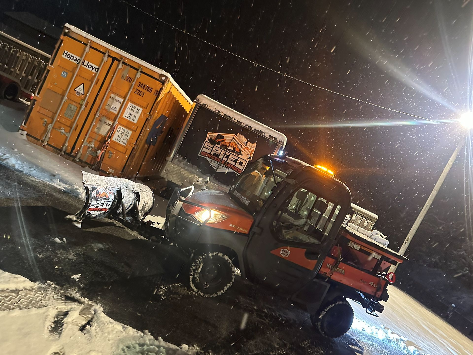 An orange utility vehicle with a snowplow parked at night near shipping containers during a snowfall.