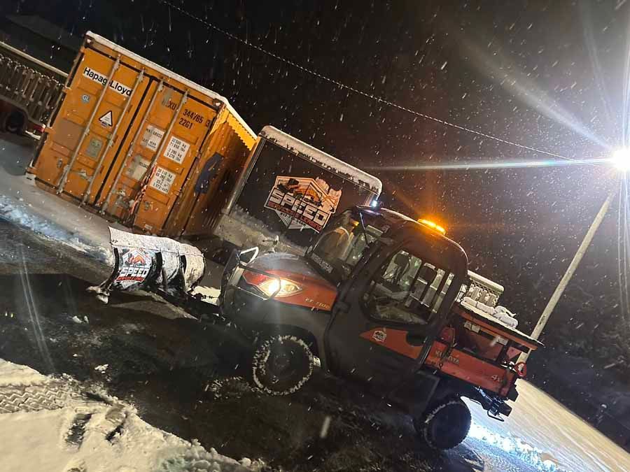 An orange utility vehicle with a snowplow parked at night in a snowy lot next to storage containers.
