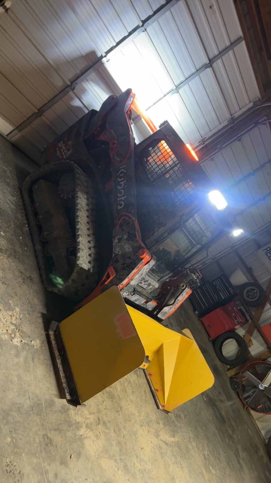 A track loader parked inside a dimly lit workshop, with a bright yellow snow blower attachment sitting on the floor.