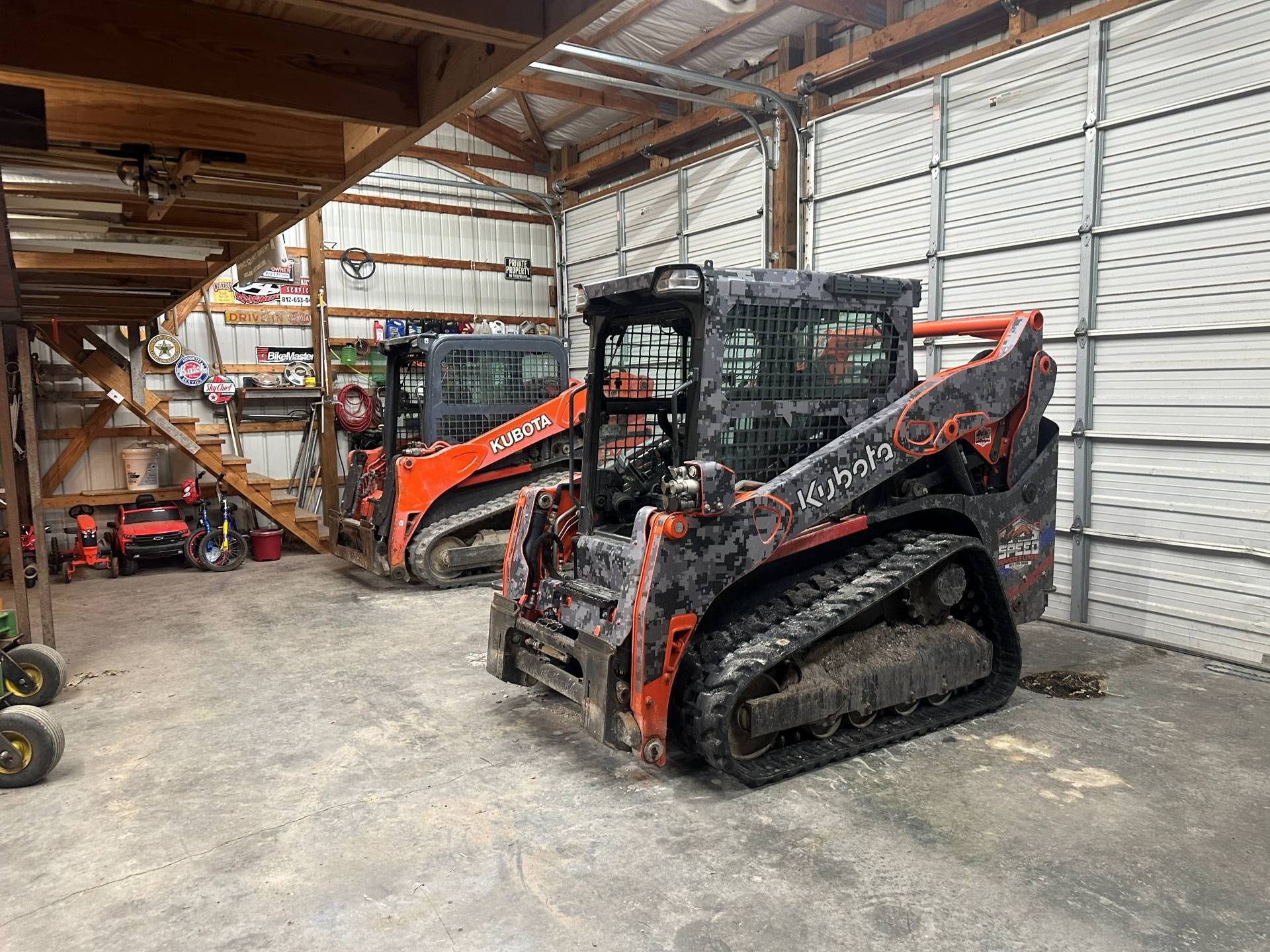Two Kubota skid steer loaders parked in a large workshop with concrete floors and metal walls.