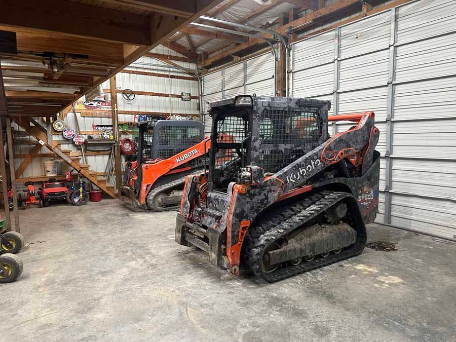 Two orange Kubota tracked skid steer loaders parked inside a spacious metal storage barn with wooden rafters.