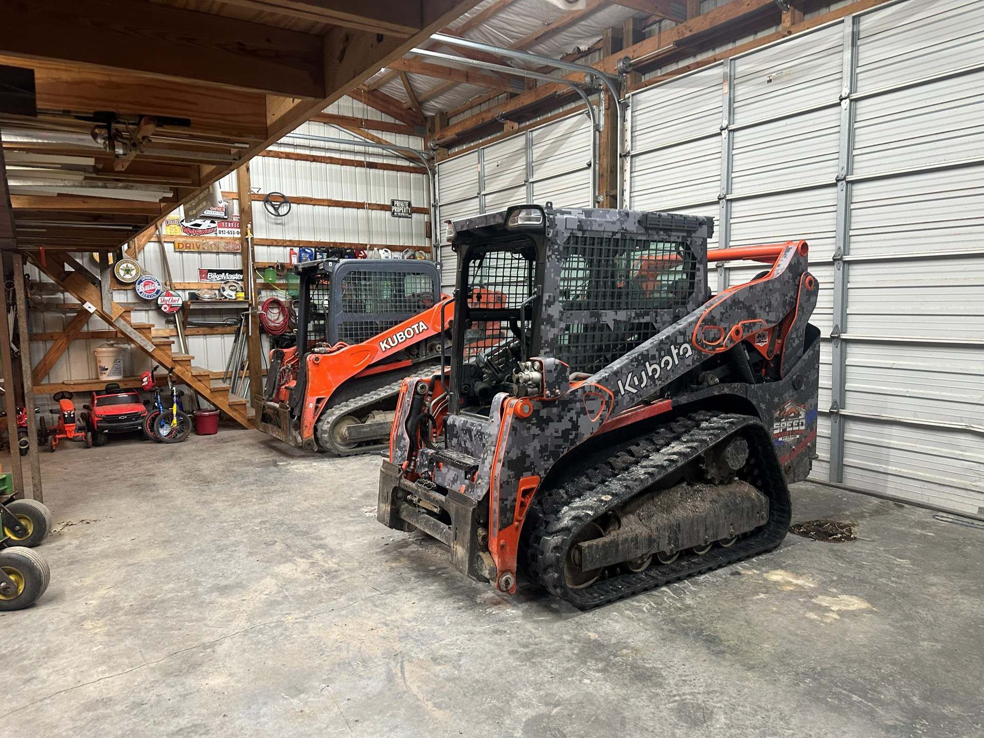 Two orange and black Kubota compact track loaders parked inside a shed with metal walls and a wooden ceiling.