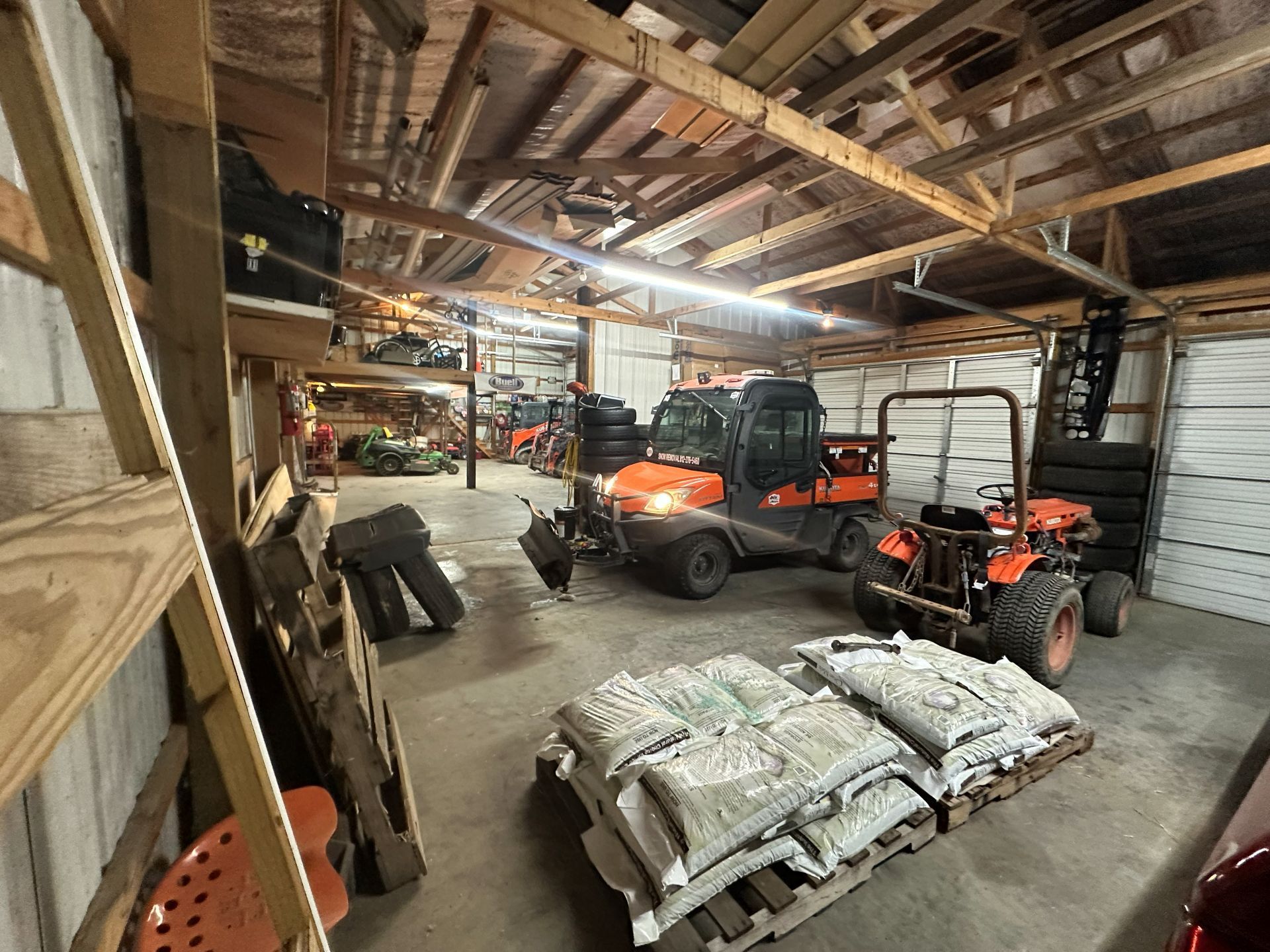 An indoor storage shed containing two orange Kubota utility vehicles, stacks of cement bags, and miscellaneous equipment.