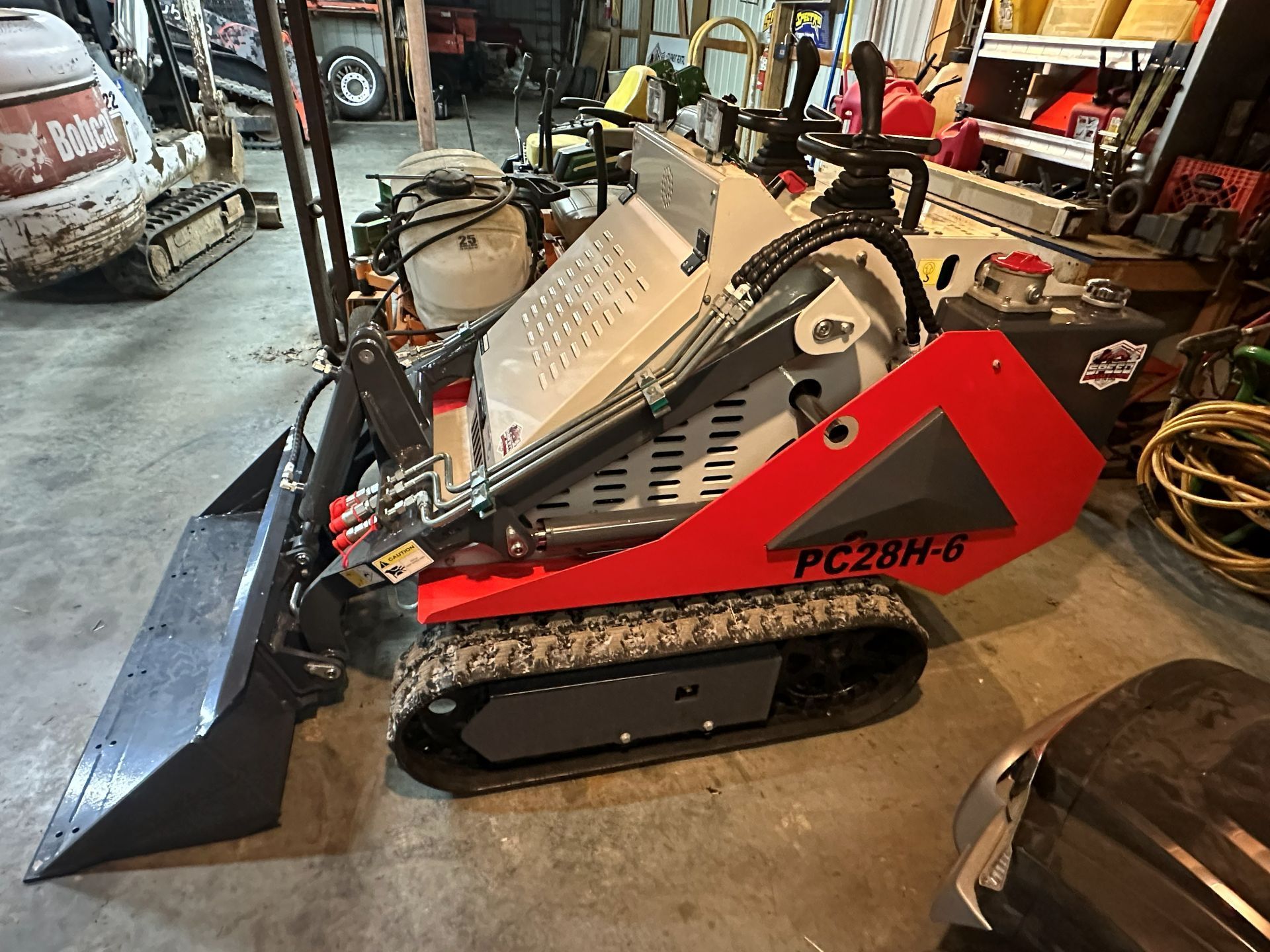 A red and white stand-on tracked skid steer loader with a front bucket parked inside a workshop.