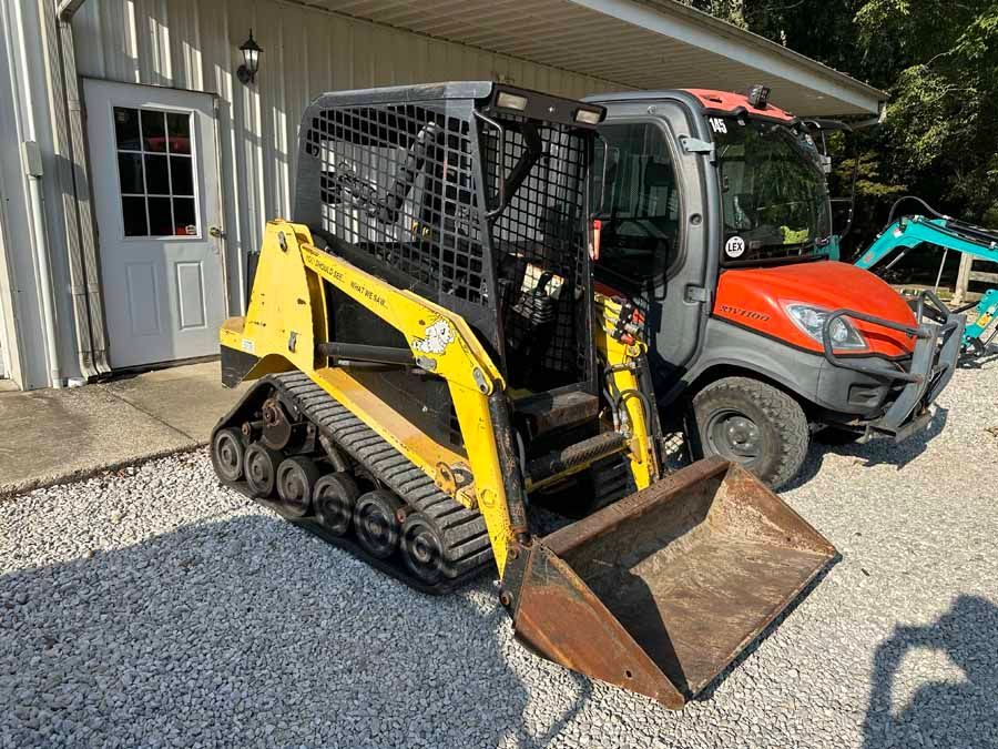 A yellow track loader with a front bucket sits on a gravel lot next to an orange skid steer and a building.