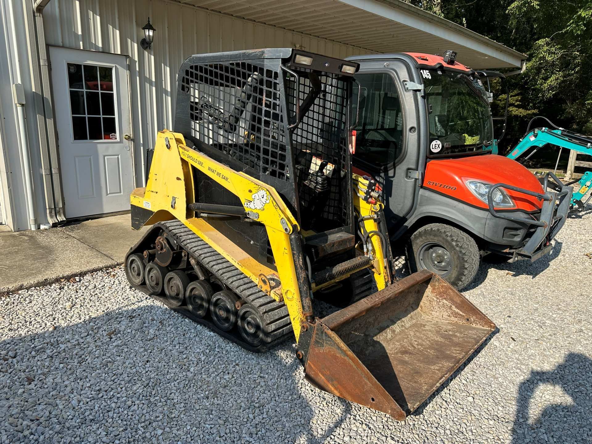 A yellow skid steer loader with tracks parked on gravel next to an orange UTV and a building entrance.