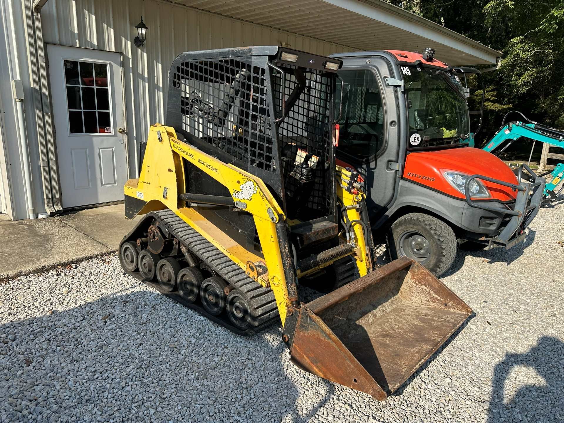 A yellow tracked skid steer loader parked on gravel next to an orange utility vehicle in front of a white building.