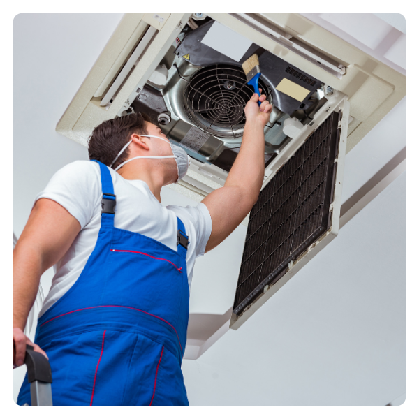 HVAC technician cleaning an air vent with a brush. White shirt, blue overalls, and mask. Inside a building.
