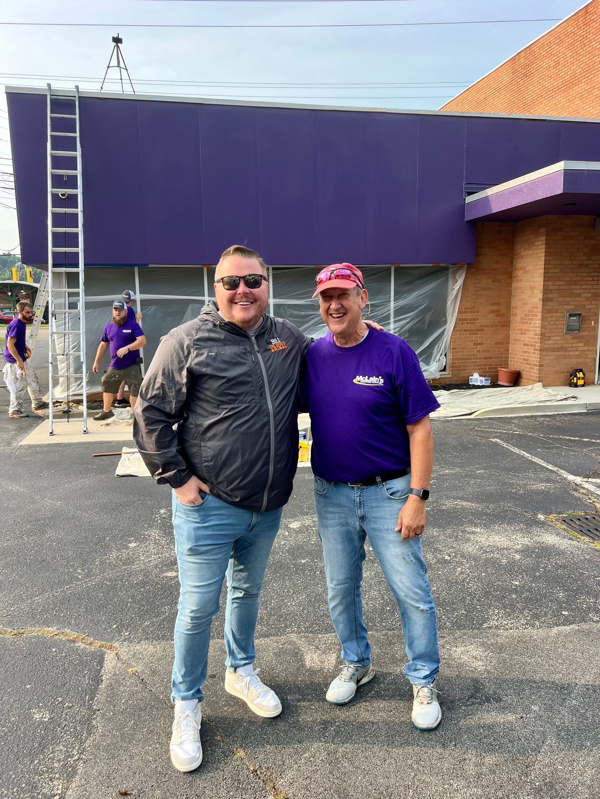 Two men are standing next to each other in front of a purple building.