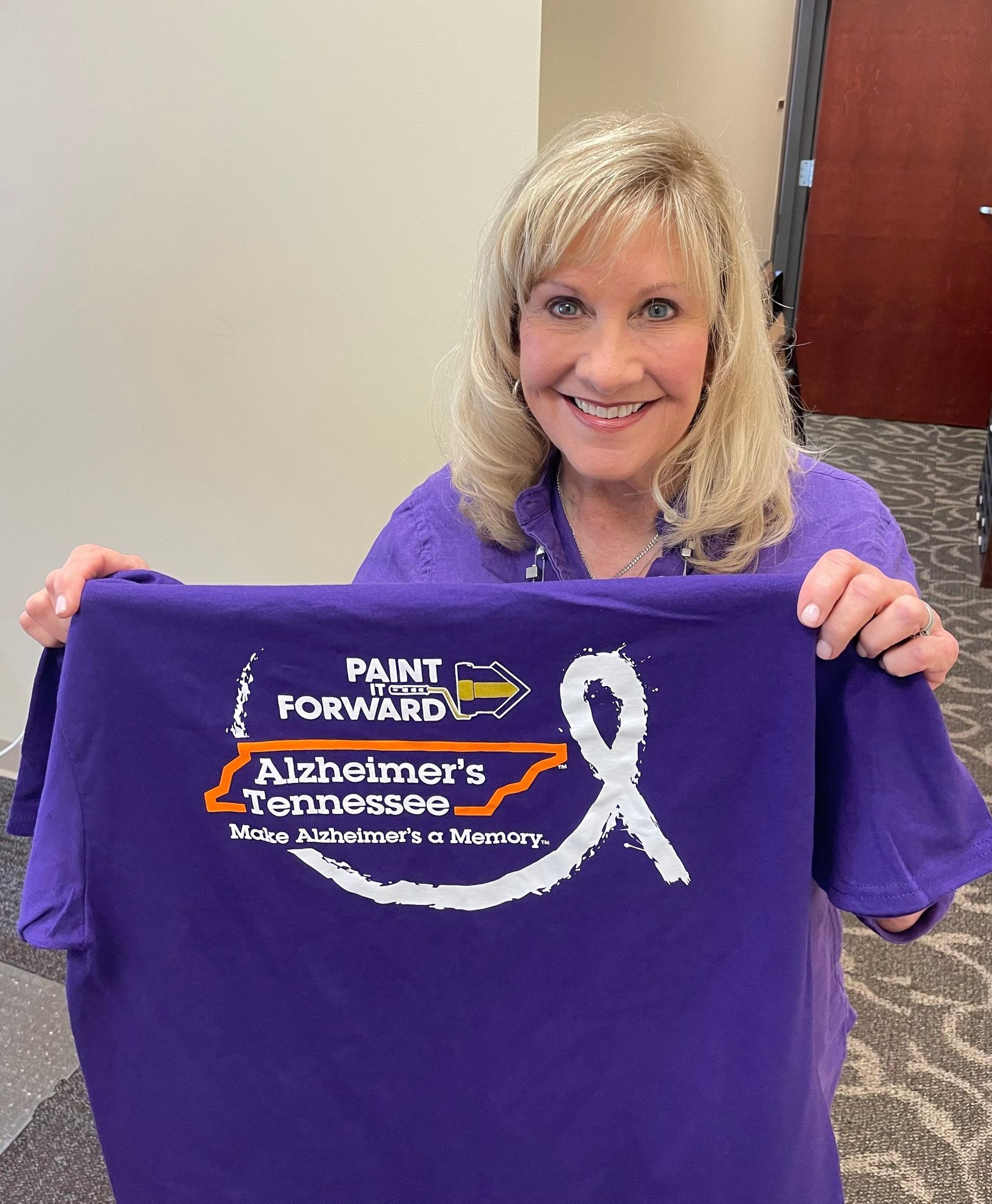 A woman is holding a purple t-shirt with a white ribbon on it.