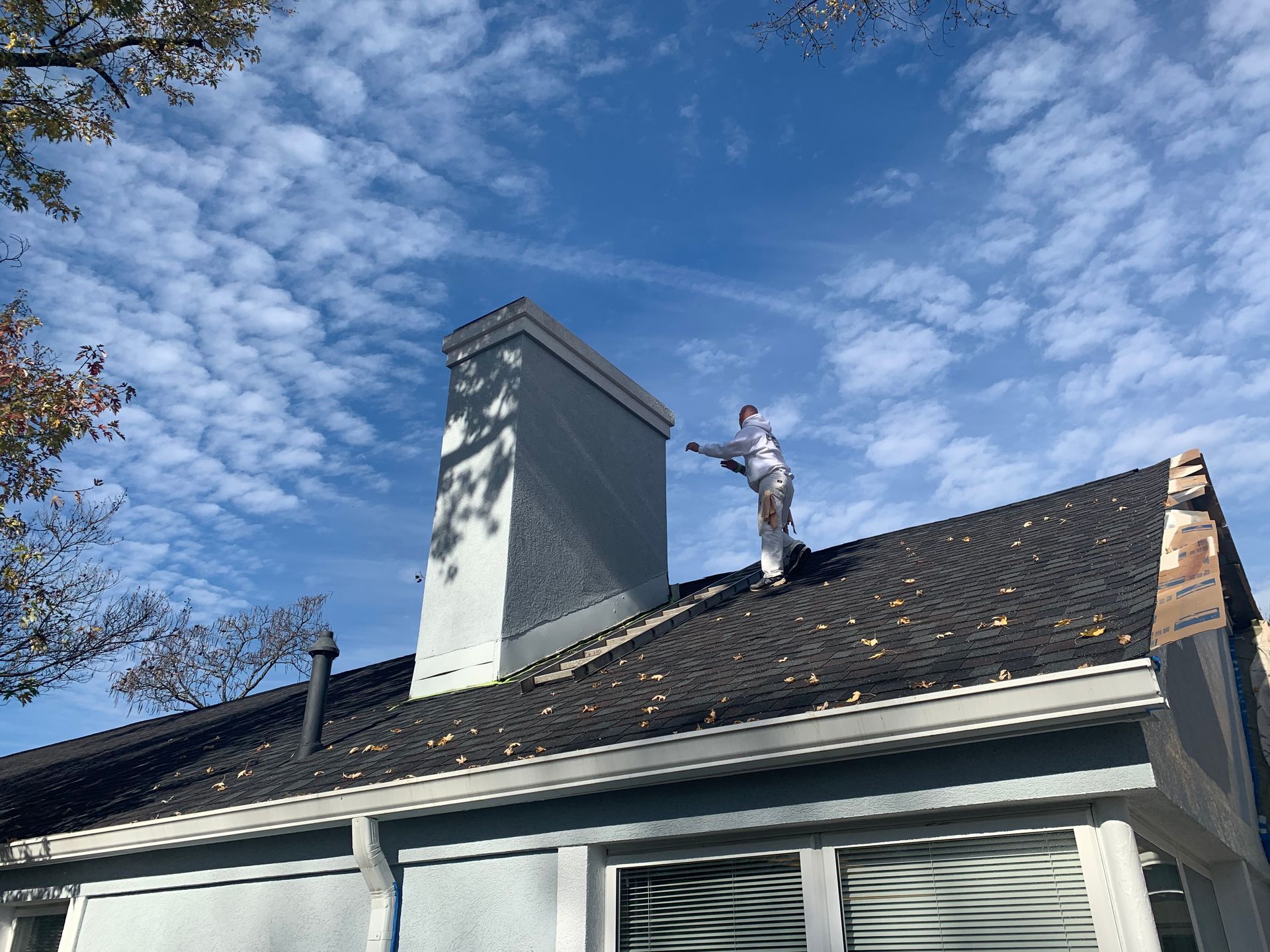 A man is standing on the roof of a house painting a chimney.