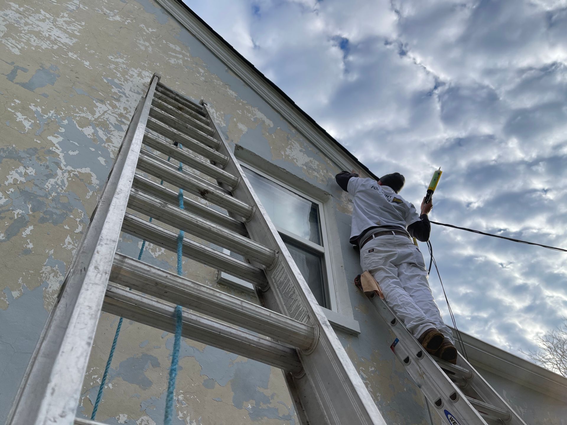 A man is standing on a ladder painting a window on the side of a building.