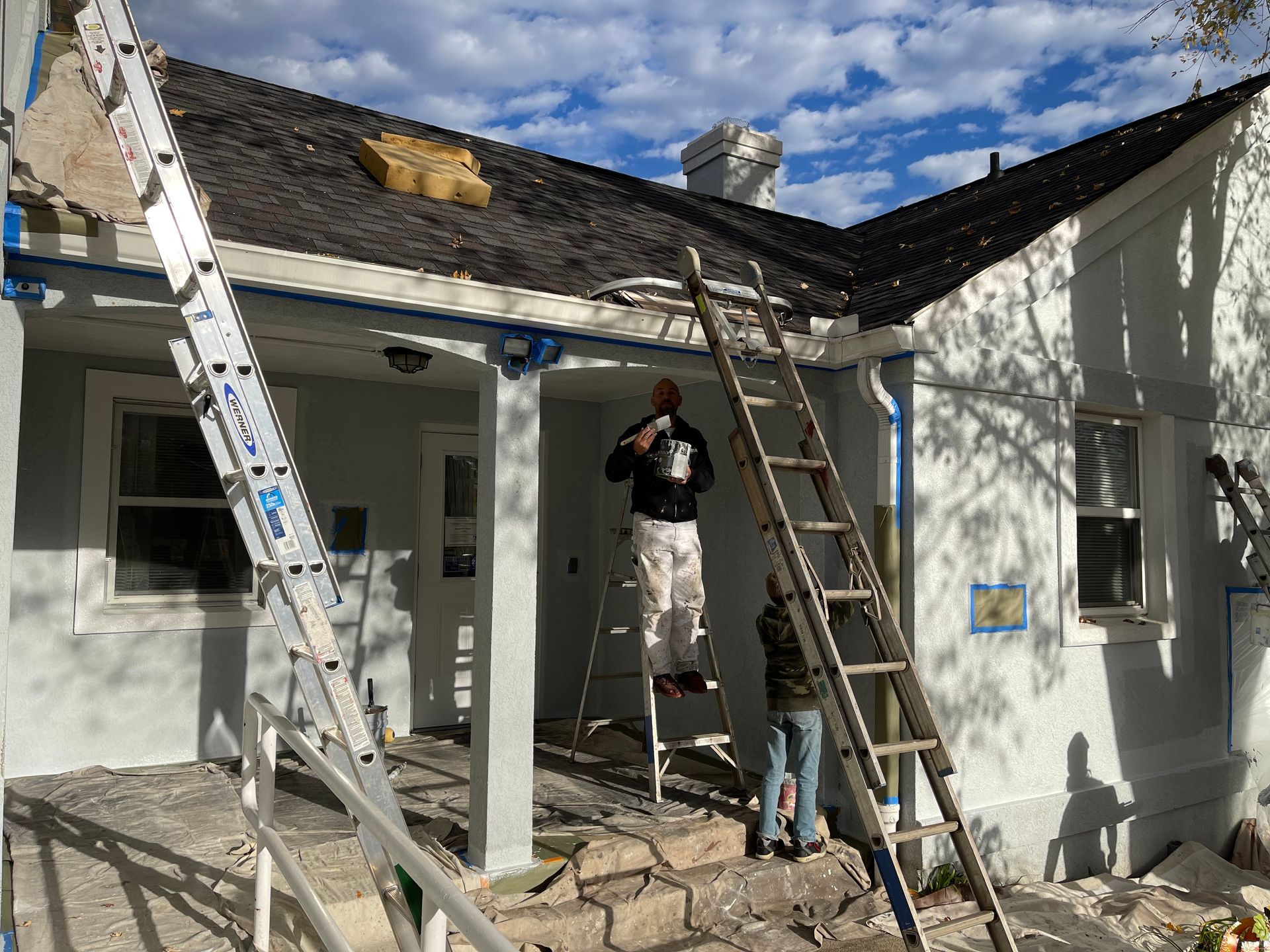 A man is standing on a ladder painting the roof of a house.