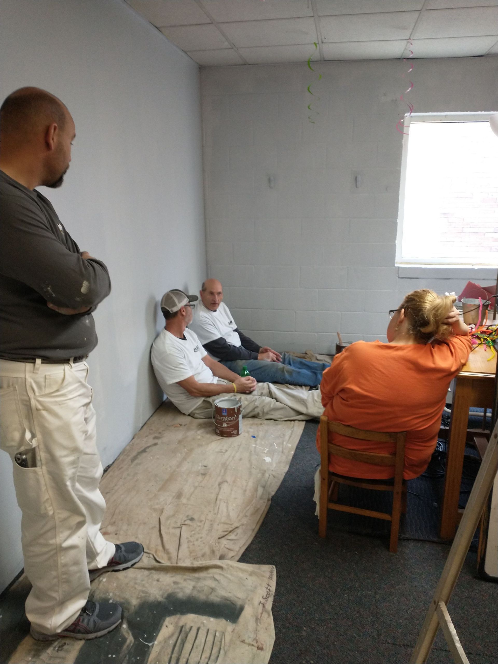 A group of people are sitting in a room painting a wall.