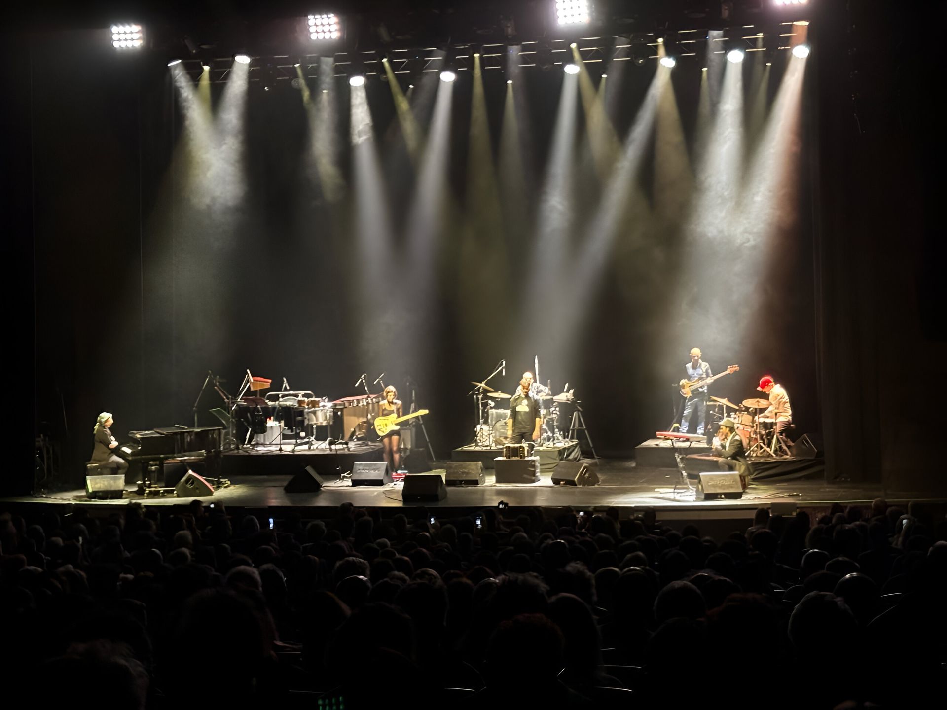 Banda tocando en el escenario, iluminada por brillantes focos. Público en primer plano, escenario a oscuras.