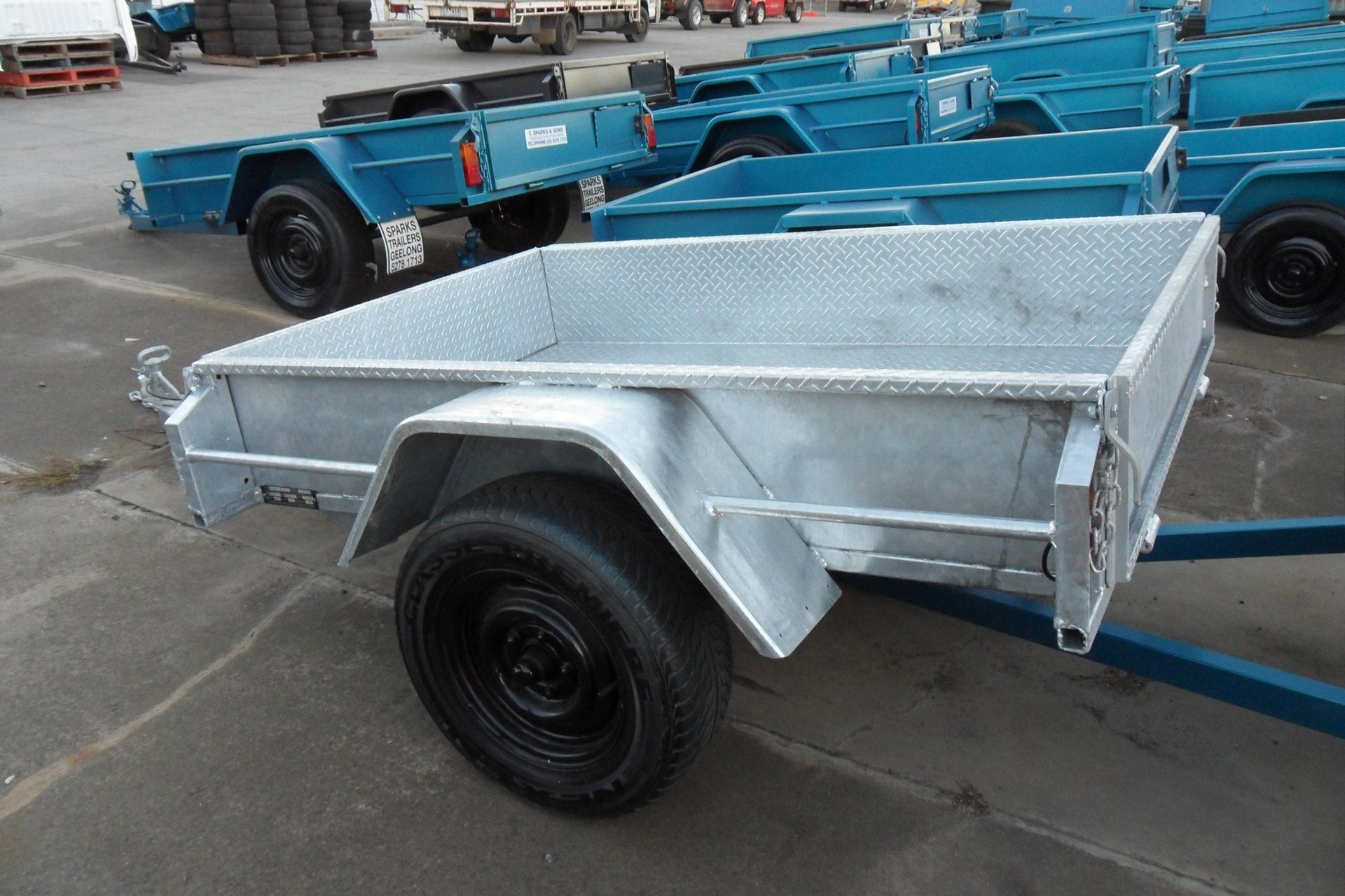 Silver utility trailer parked among blue trailers on a concrete lot