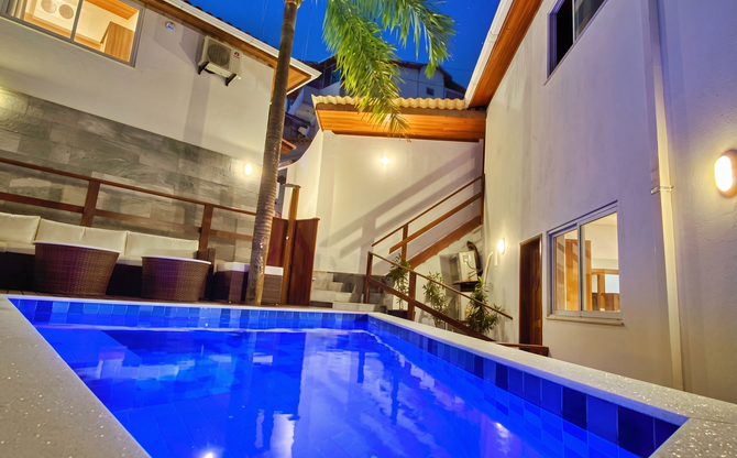 Pátio moderno com piscina iluminada em azul, paredes brancas, corrimão de madeira e luzes aconchegantes para a noite.
