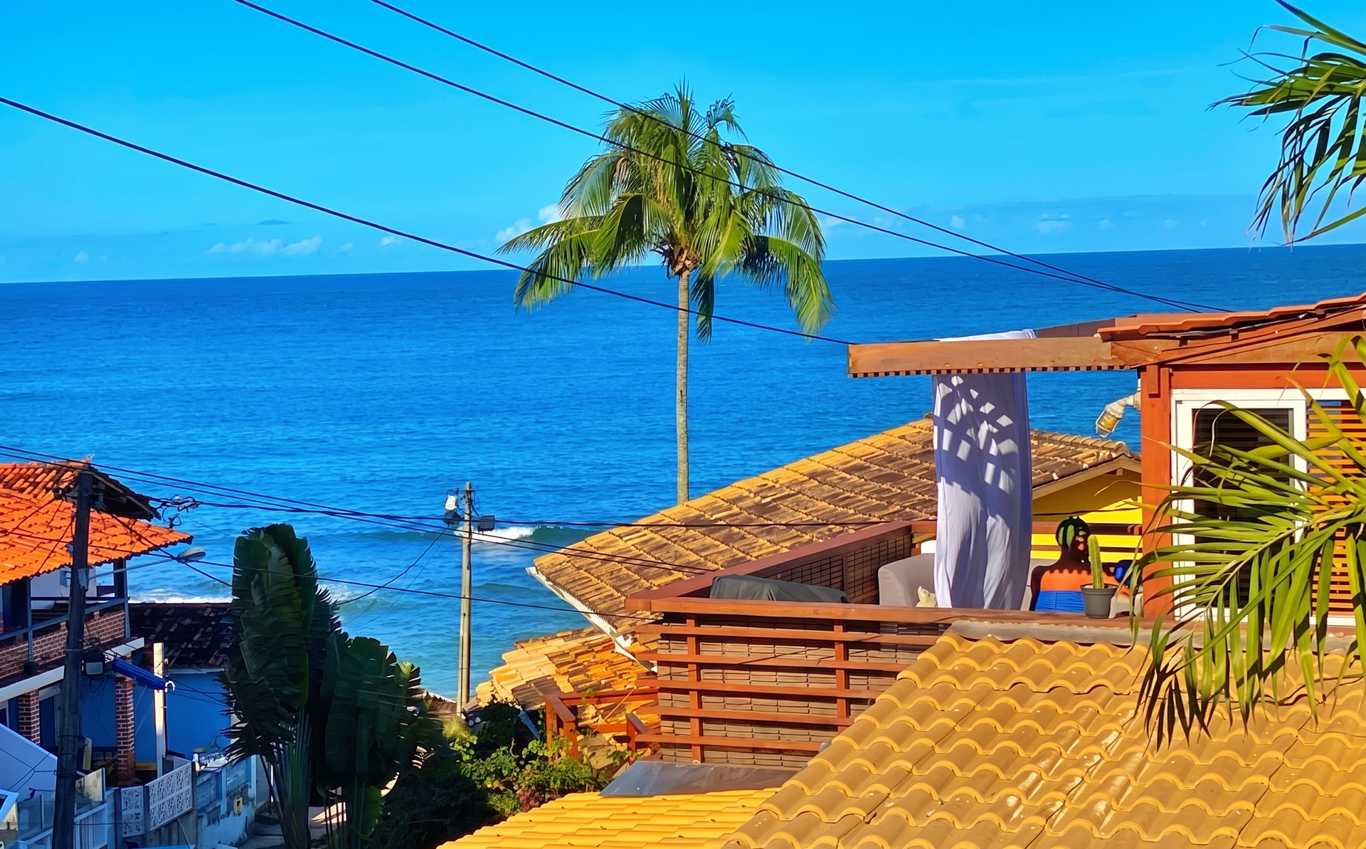 Vista tropical para o mar, com oceano azul, palmeiras, telhados de terracota e uma varanda ensolarada.