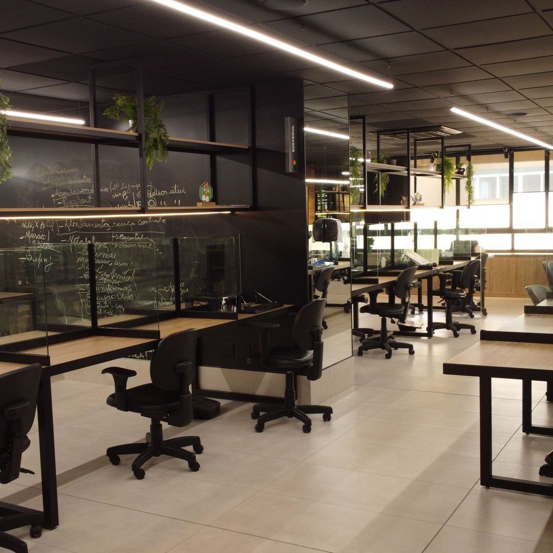 Escritório moderno com mesas pretas, cadeiras giratórias, plantas e grandes janelas em um espaço de trabalho aberto e iluminado.