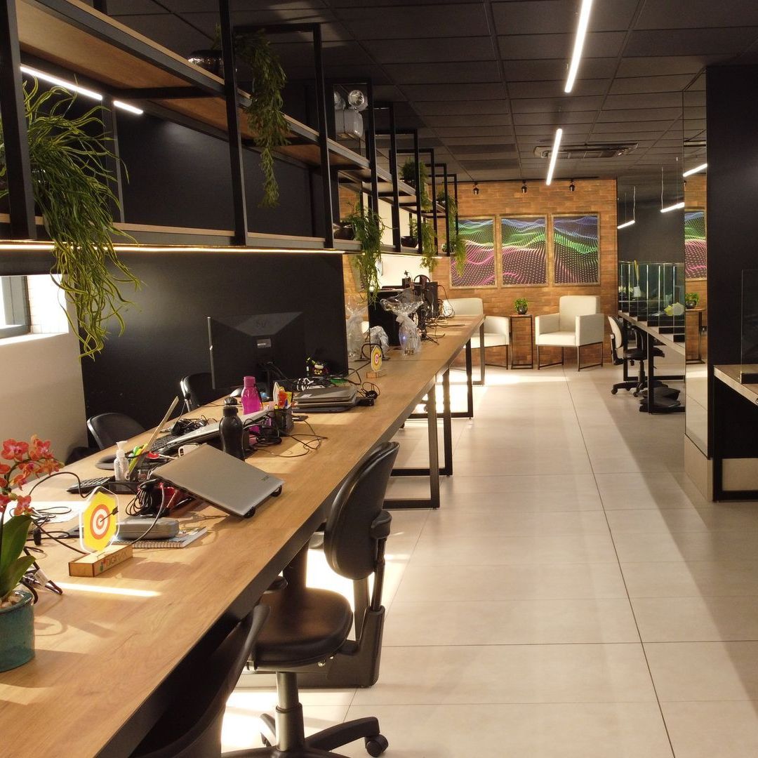 Espaço de trabalho aberto com mesas compridas, computadores, plantas e uma área de estar junto a um mural.