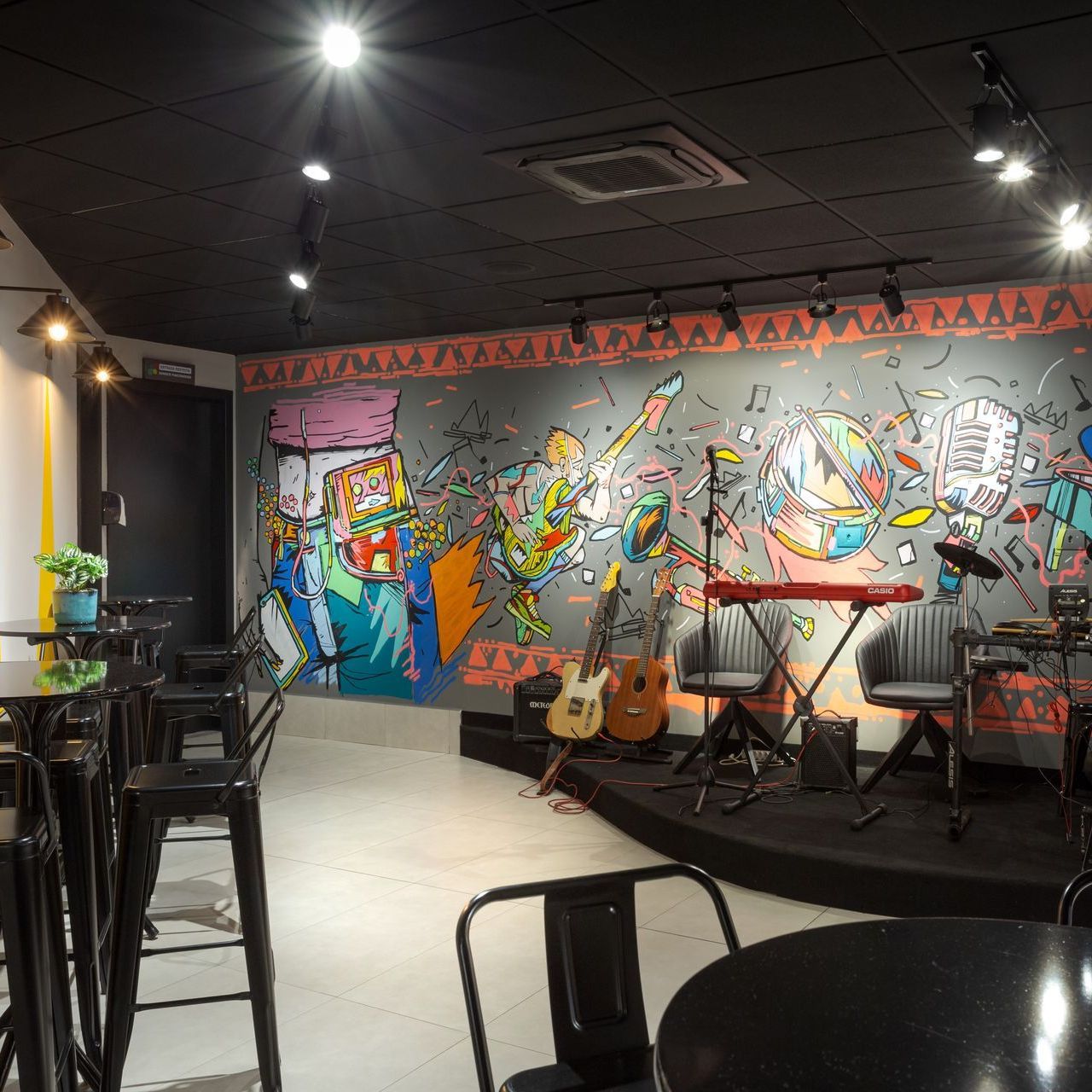 Pequeno espaço para música ao vivo com parede decorada com um mural colorido, guitarras no palco e mesas em primeiro plano.
