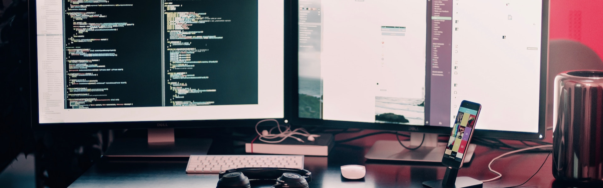 Monitor de computador exibindo código em uma tela escura ao lado de um pequeno telefone em uma mesa desarrumada.