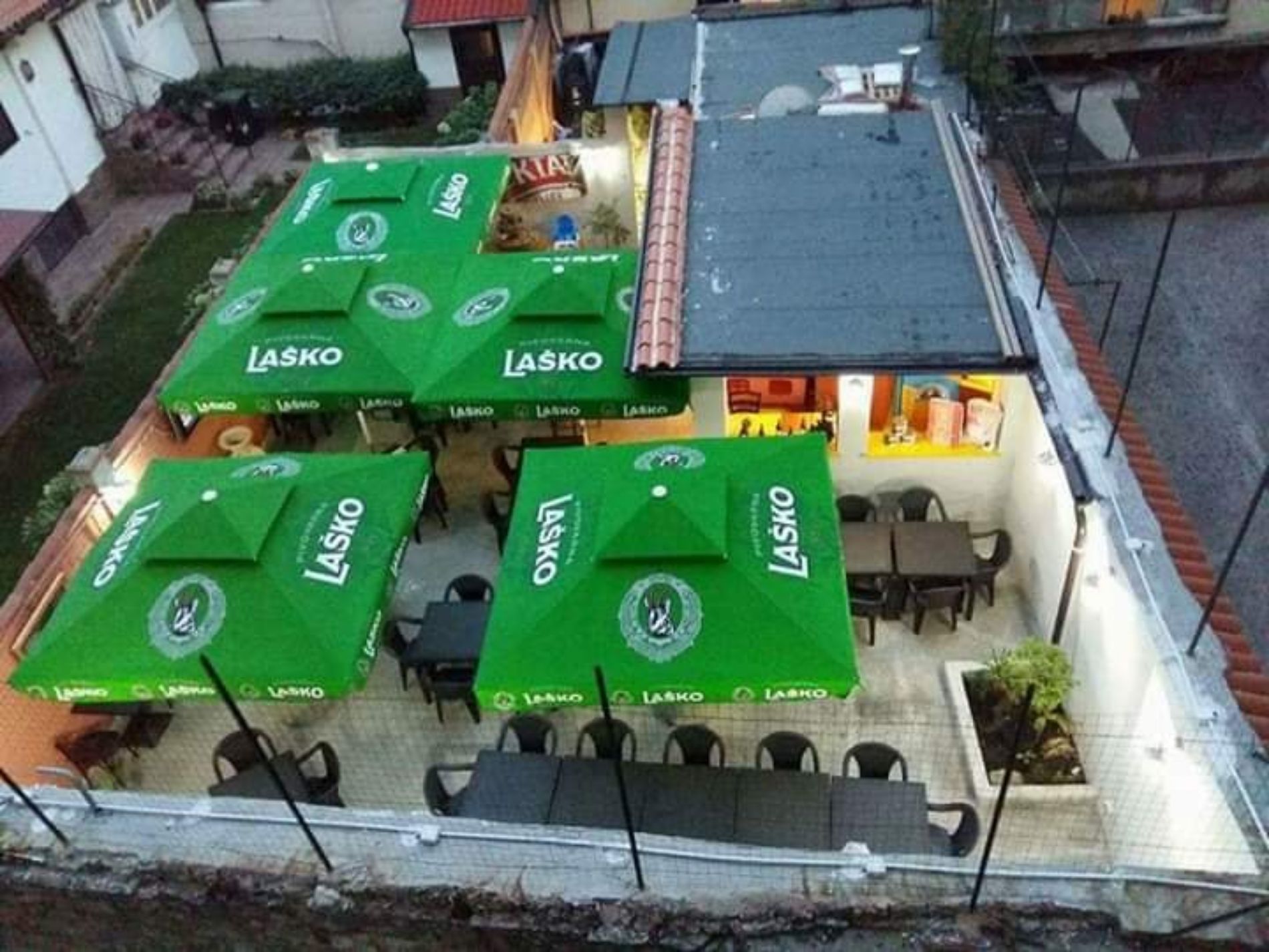 Vista aerea del patio esterno di un ristorante, con numerosi ombrelloni verdi con il marchio Laško sopra tavoli e sedie.