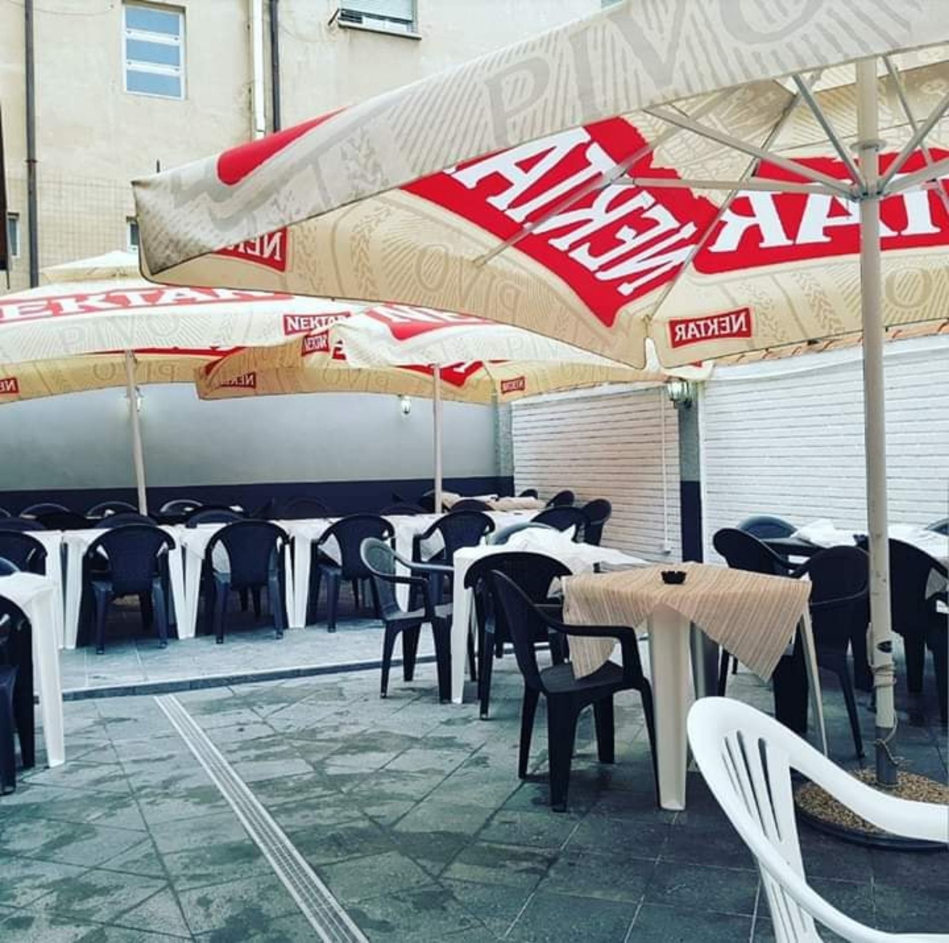 Terrazza esterna del bar con tavoli, sedie e grandi ombrelloni del marchio Nektar, pavimentata in pietra.