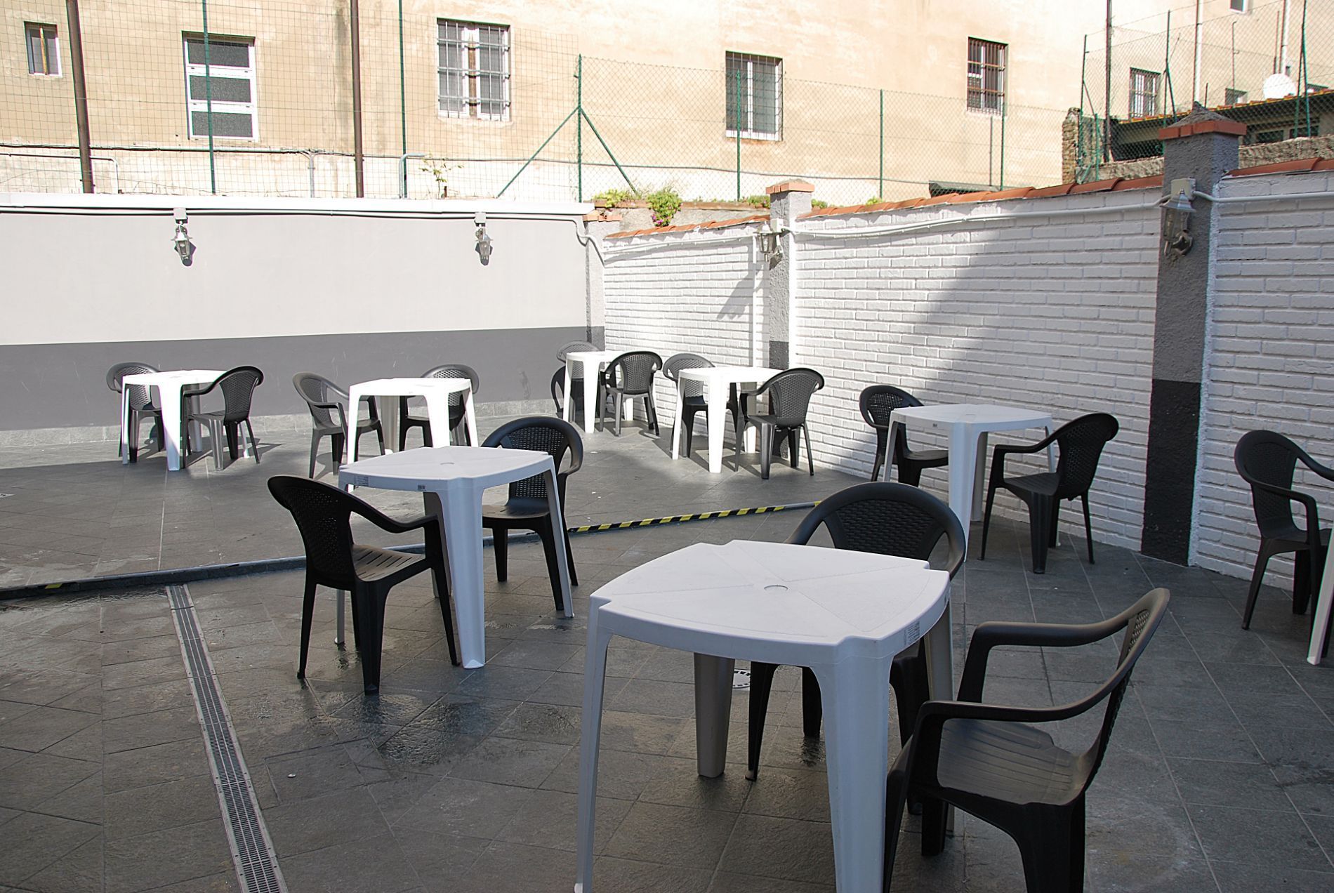 Tavoli quadrati bianchi vuoti con sedie di plastica nera disposti su un patio esterno, addossati a un muro di mattoni bianchi.