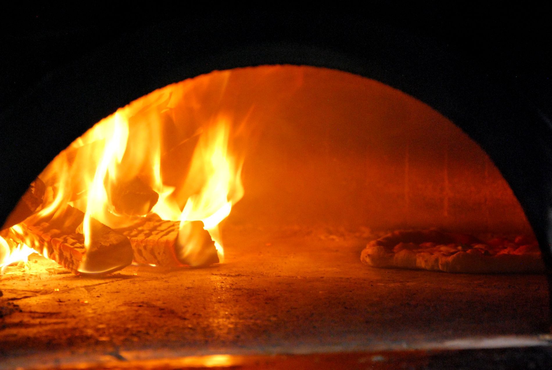 Sul lato sinistro di un forno a legna per pizza arde un fuoco vivace, mentre sulla destra una pizza sta cuocendo sul piano rovente.