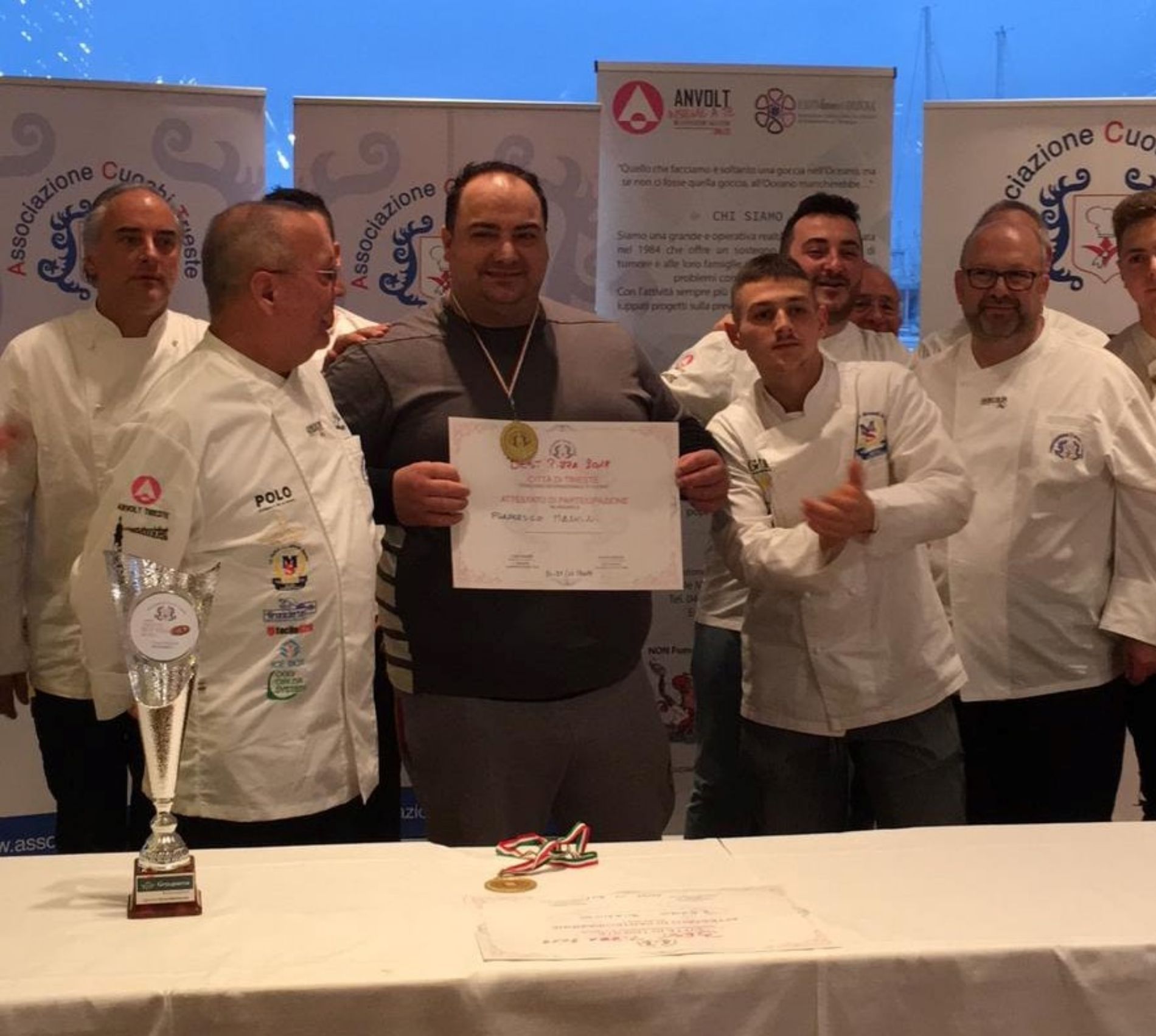 Un gruppo di chef in uniforme bianca circonda una persona che regge un attestato di premiazione durante un evento culinario.