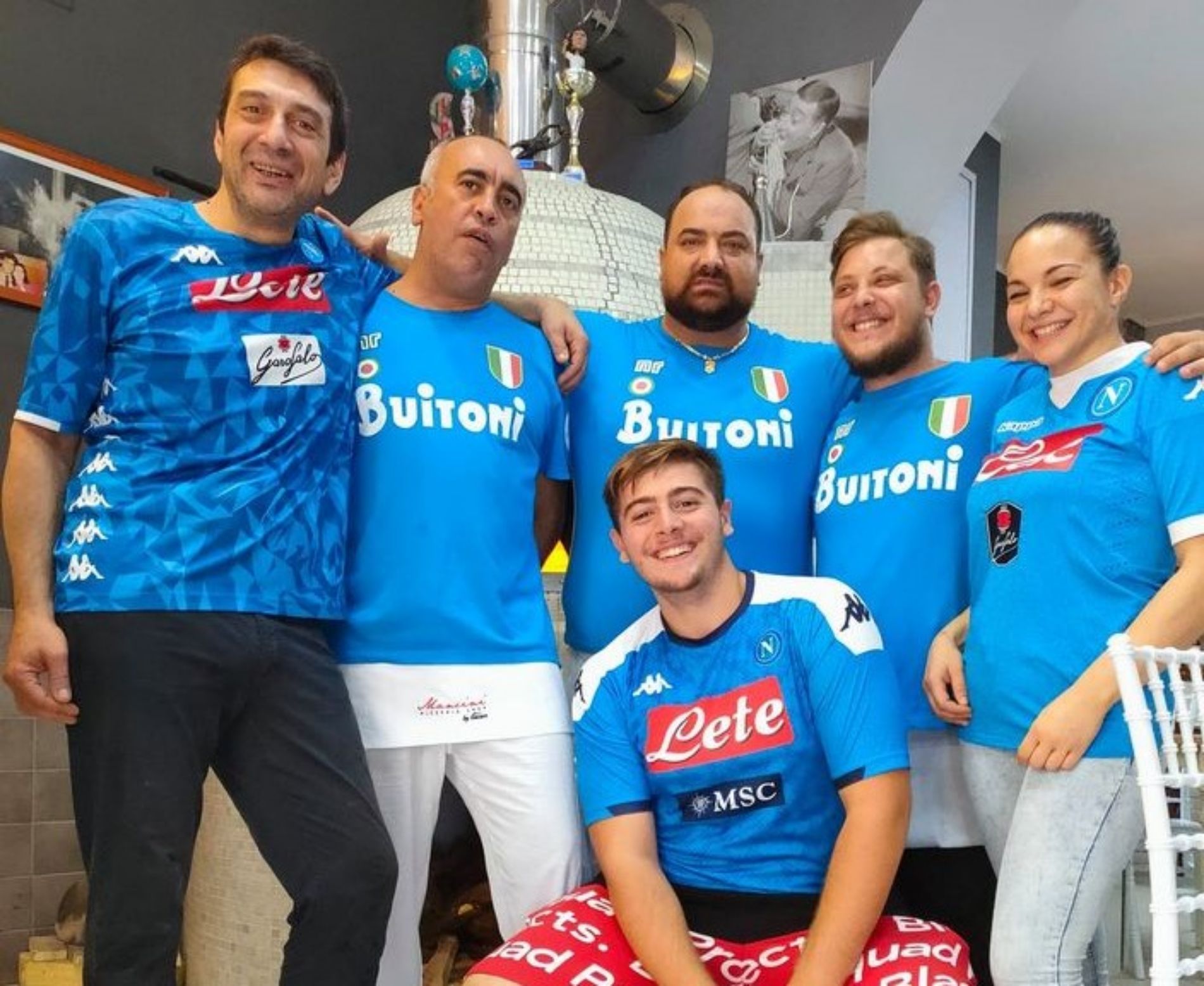 Un gruppo di sei persone che indossano maglie blu del Napoli sorride per una foto in interni, una delle quali seduta in prima fila.