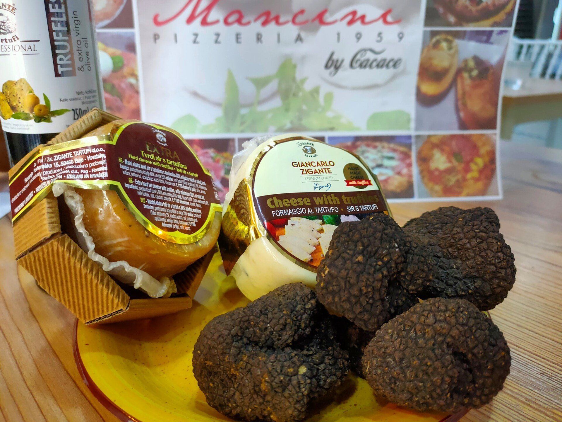 Tre tartufi neri interi sono adagiati su un piatto giallo, davanti a due forme di formaggio aromatizzato al tartufo e a una bottiglia d'olio.