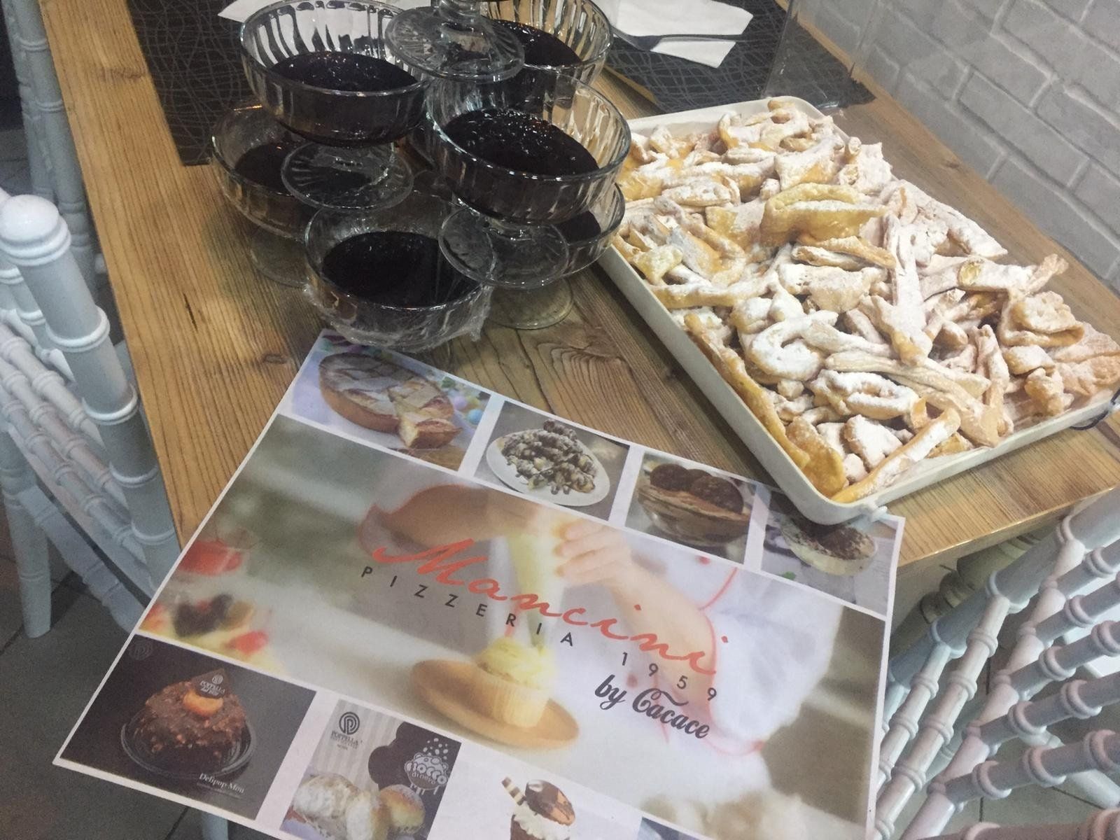 Un vassoio di pasticcini fritti cosparsi di zucchero, ciotole di vetro con salsa scura e un menù della pasticceria su un tavolo di legno.