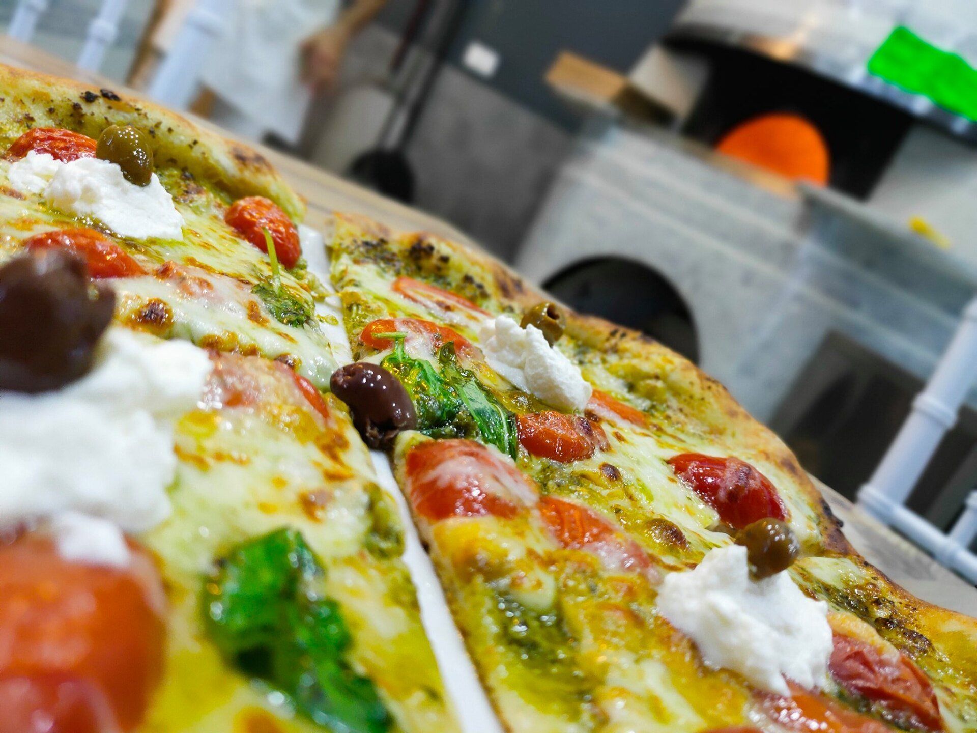 Primo piano di una pizza condita con pesto, pomodorini, olive e fiocchi di formaggio bianco, con un forno a legna sullo sfondo.