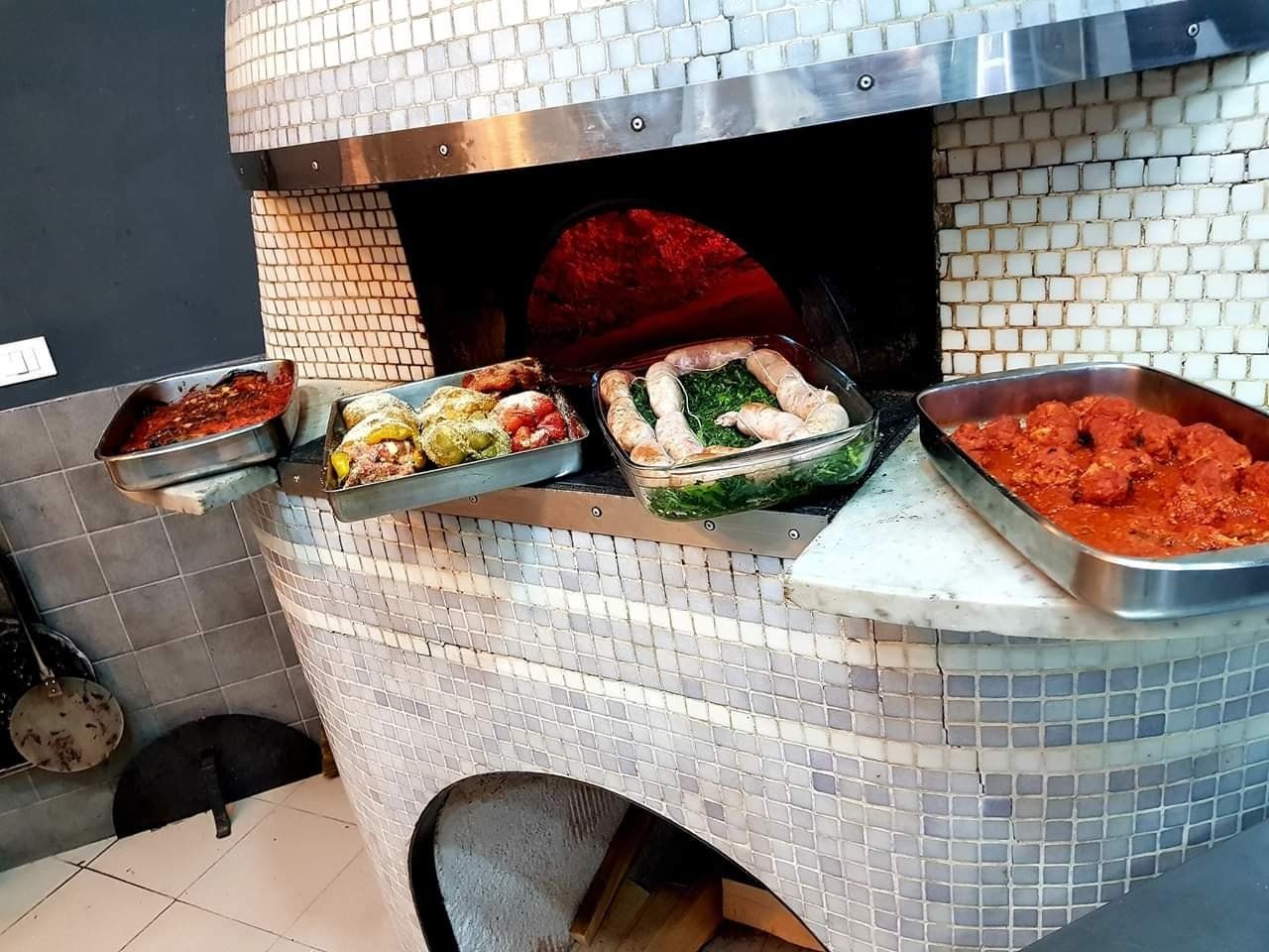 Quattro vassoi di metallo contenenti cibo sono appoggiati sul bordo di un forno per pizza in mattoni piastrellati.