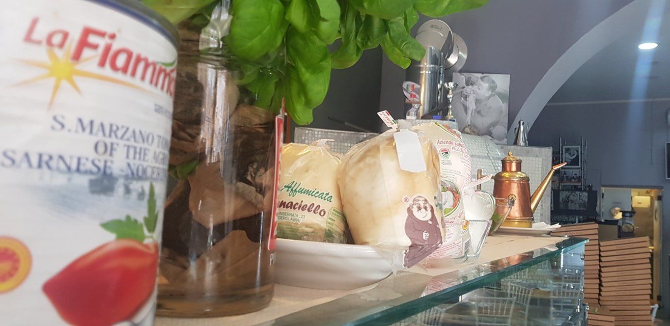 Un primo piano del bancone di un ristorante con una lattina di pomodori La Fiammante, basilico fresco e scatole di pizza impilate.